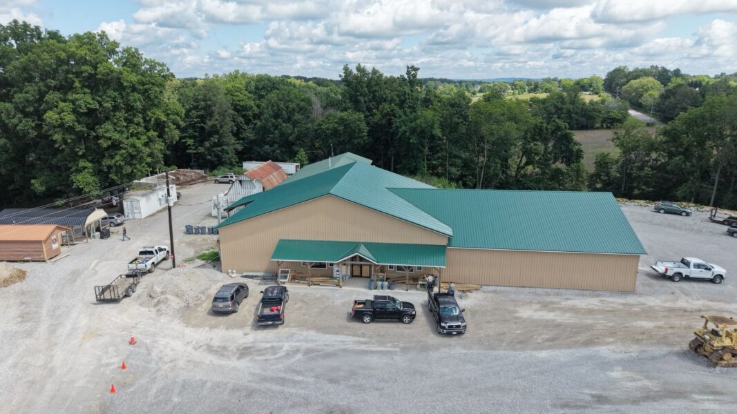 Aerial view of a large commercial building with a green metal roof, surrounded by gravel parking and various vehicles. The site features additional storage structures and is nestled among trees, indicating a rural setting. Ideal for showcasing business facilities or outdoor services.