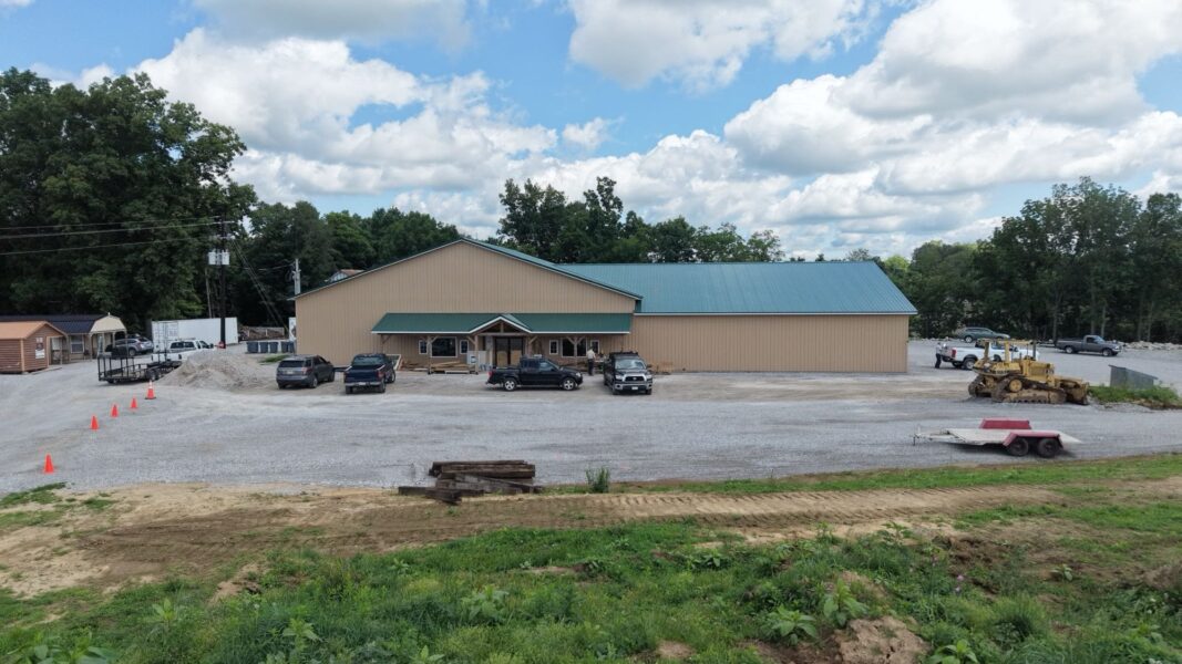 Large commercial building with a green metal roof and beige exterior, surrounded by gravel parking and orange traffic cones. Various vehicles are parked in the lot, with construction equipment visible in the background. Lush greenery and a cloudy sky complete the scene.